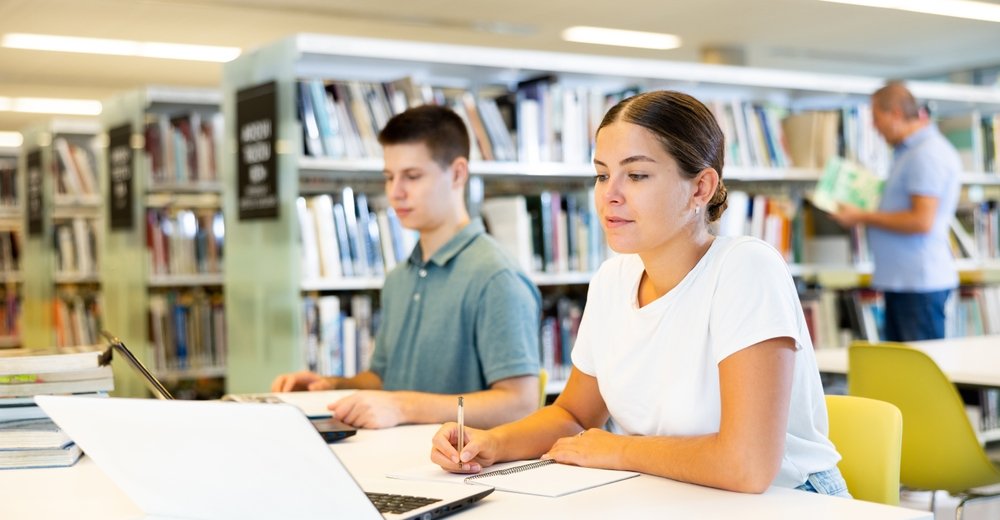 Two students studying in a library, one taking notes in a notebook and the other using a laptop, with bookshelves in the background.